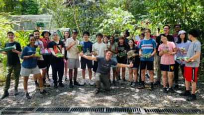 A group of people of various ages stand outside on a gravel path in lush greenery, holding plants and smiling at the camera.