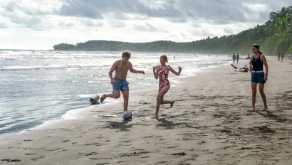 People playing soccer on a sandy beach with the ocean and distant greenery under a cloudy sky.