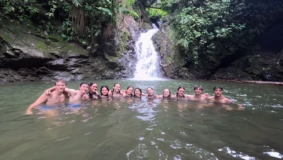 A group of people swimming in a natural pool at the base of a small waterfall, surrounded by lush greenery.