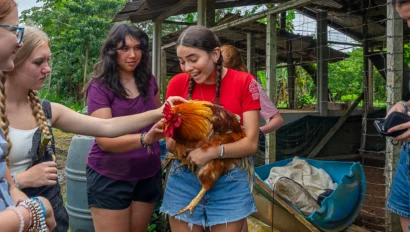 A group of young people gather around a person holding a large rooster, with others gently petting it, near a rustic outdoor shelter.