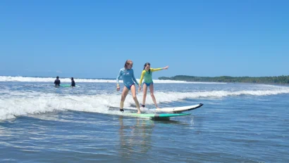 Two people surfing on small waves, wearing blue and yellow rash guards, with a clear sky background.