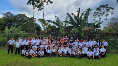 A large group photo of students and teachers standing and sitting on grass, with trees and a fence in the background.