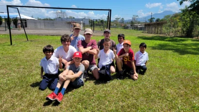A group of children and two adults pose on a grassy field with a soccer goal in the background under a clear blue sky.