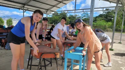 Four young people clean and sand desks under a metal-roofed outdoor shelter on a sunny day.
