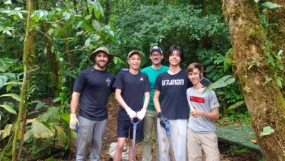 Five people with gardening tools stand together in a lush forest setting. They are smiling and appear to be engaged in outdoor work or conservation efforts.