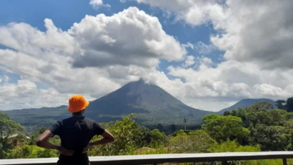 Person in an orange hat stands on a balcony, looking at a large volcano under a partly cloudy sky. Lush greenery surrounds the area.