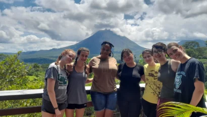 Seven people stand together on a balcony with a scenic view of a large, conical mountain in the background under a partly cloudy sky.
