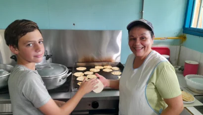 A person and a woman are making tortillas on a griddle in a kitchen. The woman is smiling, and both are wearing casual clothes.