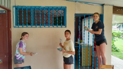 Three people painting a beige wall outside a building with blue window and door frames. Two stand on the ground, while one is on a ladder. They are holding paintbrushes and small trays.