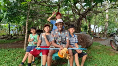 A group of five smiling boys are sitting on a wooden bench under a tree. The central boy holds gardening tools. They are outdoors in a grassy area with a scooter nearby.