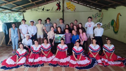 A group photo of people indoors, with women in red and white dresses kneeling in front and others standing behind them under a wooden roof. Wall decorations include music notes and masks.