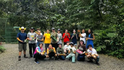 A group of people stands outdoors holding various items, surrounded by lush green trees and plants.