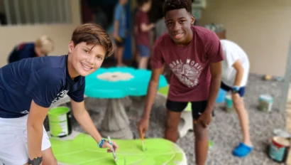 Two boys are painting a bench green outdoors. Other people are blurred in the background.
