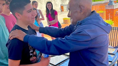 An older man places his hands on a teenage boy’s shoulders during a civil rights school trip, while others stand nearby under a metal canopy in an outdoor setting.