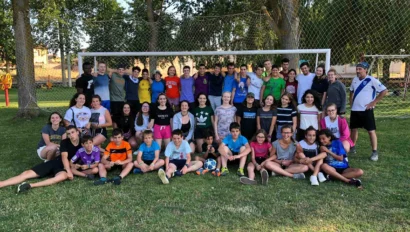 A group of people, including adults and children, pose in front of a soccer goal on a grassy field with trees in the background.