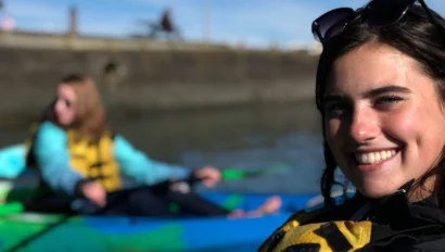 Two people kayaking; one in focus smiling, wearing sunglasses and a life jacket, and the other blurred in the background.