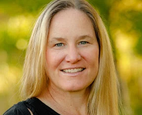 Woman with long blonde hair smiling outdoors with blurred green background.
