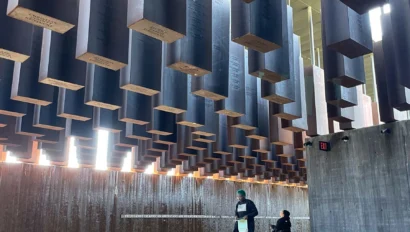 Individuals sit and stand on tiered steps under numerous suspended rectangular blocks in a sunlit, modern architectural space.