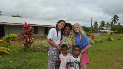 Five people, including three adults and two children, stand together outside on grass with buildings and palm trees in the background, smiling at the camera during a Fiji student trip.