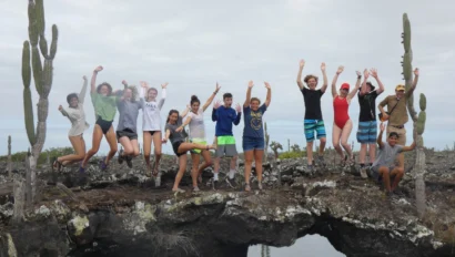 A group of people jumps in unison on a rocky bridge surrounded by cacti and water.