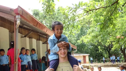 A child wearing a blue shirt sits on a man's shoulders outside, surrounded by other children in a schoolyard. Trees and a building are in the background.