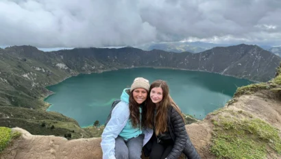 Two women, part of a teen travel program, sit on a rocky ledge overlooking a vast crater lake surrounded by mountains under a cloudy sky.