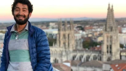 A person with a beard smiles at the camera while standing in front of a blurred cityscape and cathedral at sunset.