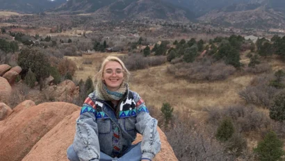 A person sits on a rocky outcrop wearing a colorful jacket, with a mountainous landscape in the background under a cloudy sky.