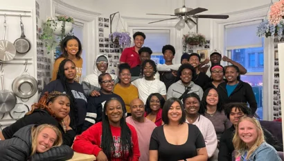 A group of people gathered in a cozy kitchen, smiling at the camera. Pots and pans hang on the wall.