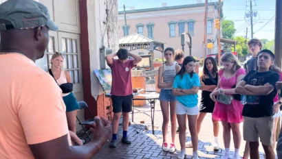 A man speaks to a group of attentive teenagers gathered on a brick sidewalk in a town setting, with buildings and street signs visible in the background.