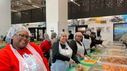 A group of people wearing hairnets and aprons prepare food at a counter in a bright kitchen space.