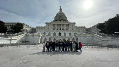 A group of people stands in front of the United States Capitol building with a cloudy sky and sun visible in the background.
