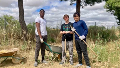 Three students stand outdoors on dry ground, holding gardening tools and wearing gloves, with a wheelbarrow and tall grass behind them—capturing a moment from a Spain school trip under a partly cloudy sky.