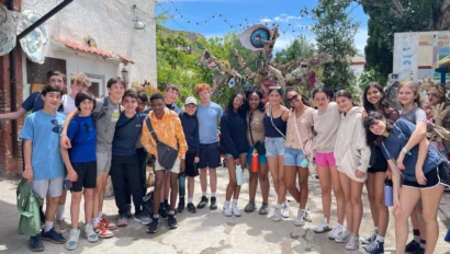 A diverse group of teenagers stand together outdoors, smiling in front of an artistic sculpture made from various materials. Trees and a building are visible in the background.