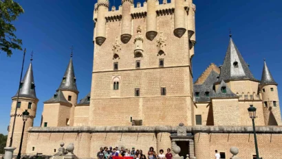 Group of people holding a Spanish flag in front of a historic castle with tall, ornate towers and a clear blue sky.