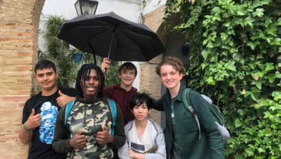 Five people standing in front of a brick archway with greenery. One holds an umbrella. Some are smiling and giving a thumbs up.