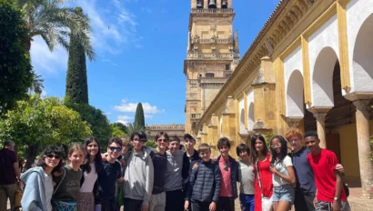 A group of people pose for a photo in front of a historic bell tower and arches, with a blue sky and palm tree in the background.