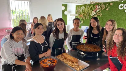 Seven people are standing around a table, smiling. The table has a large pan of food, a punch bowl with fruit, and various snacks. They are in a room with floral decor and a green wall.