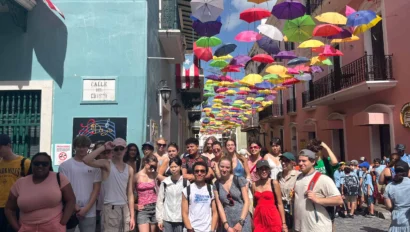 A group of people pose on a colorful street with hanging umbrellas in a sunny setting.