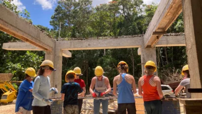 A group of people wearing hard hats and safety gear work together on a construction project at an outdoor site surrounded by trees.