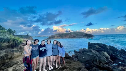 Six people stand together on rocky terrain by the ocean under a partly cloudy sky.