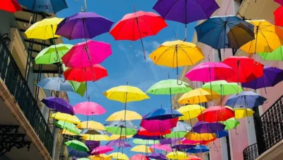 Colorful umbrellas hang across a narrow street between pastel buildings under a clear blue sky.