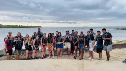 A group of people in life jackets posing for a photo on a beach with water and cloudy sky in the background.
