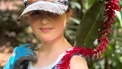 A person in a colorful cap stands near red tropical flowers in a lush, green forest, wearing a patterned shirt and a blue glove.