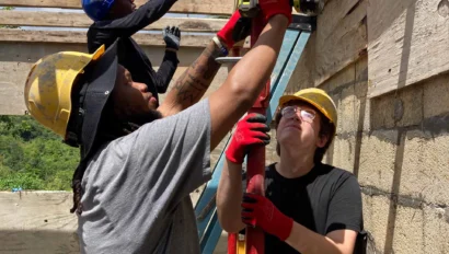 Three construction workers wearing helmets and gloves are working together to secure wooden beams on a building site under a partly cloudy sky.