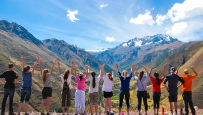 A group of people facing away, raising their arms, overlooks a mountainous landscape with a bright blue sky.
