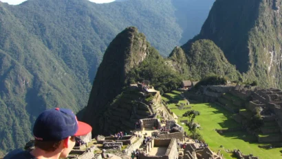 Person with a hat overlooking Machu Picchu ruins, surrounded by mountains under a clear sky.