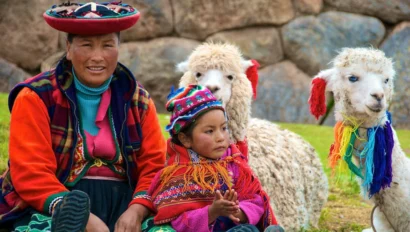 A woman and child in colorful traditional clothing sit on the grass with two alpacas, in front of a stone wall.
