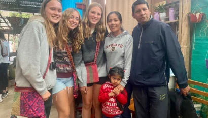A group of six people, including four young women, one older woman, and a man, are posing together indoors. A child stands in front, wearing a red sweater.