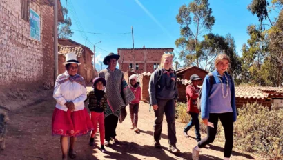 A group of people, including children and adults, walk along a dirt path in a rural village setting, with brick buildings and trees in the background under a clear blue sky.